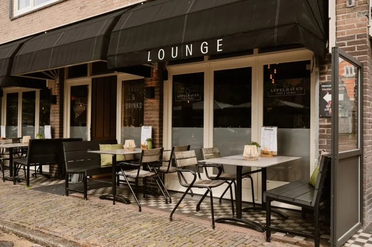 Een buitenterras van een restaurant met de naam "Appelhaven Food & Drinks," voorzien van zwarte luifels met de tekst "Lounge," houten tafels en stoelen, menu's op de tafels en een geplaveide straat.